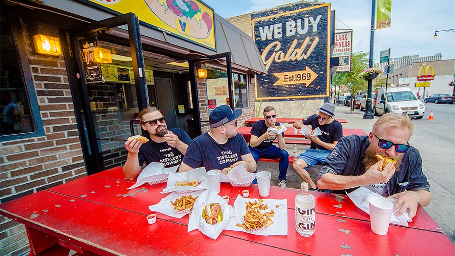 group of five young adult males sit at red benches eating Chicago-style hots dogs with french fries