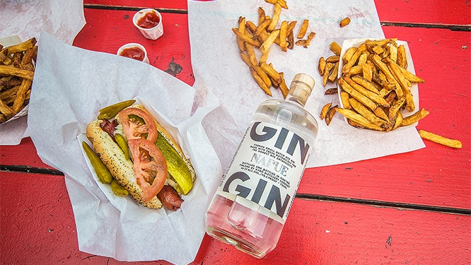 bottle of gin on a red tabletop with Chicago-style hot dogs and french fries