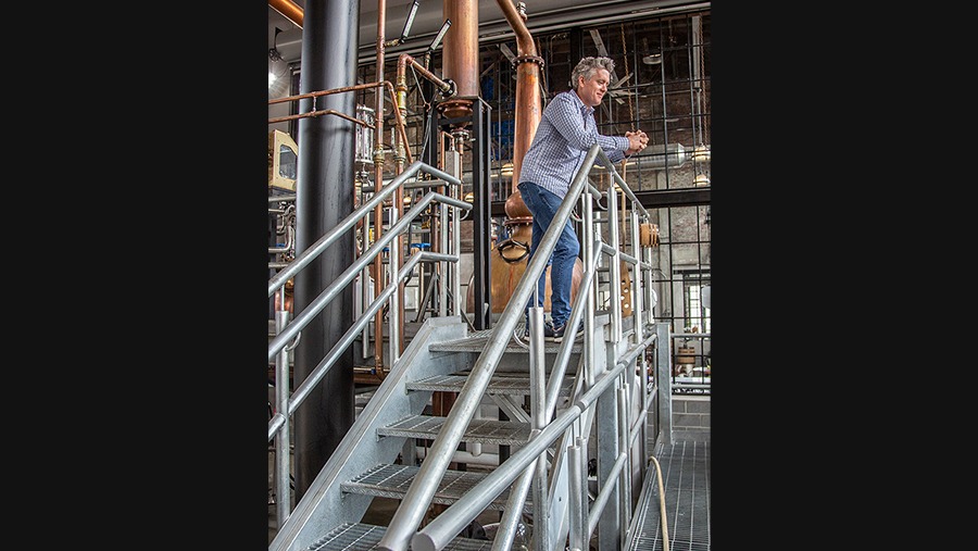 grey hair man in flannel shirt leaning against metal railing in front of copper stills and pipes