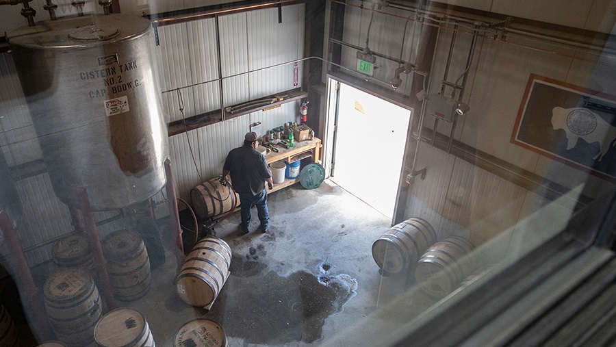 looking down on man in a garage filling wooden barrels with whiskey from a large steel vat on stilts