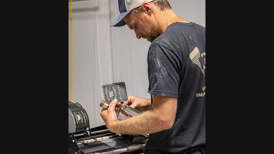 Head distiller Sam Mead, a fair skin man in a baseball cap and neutral t-shirt, applying labels to glass bottles 