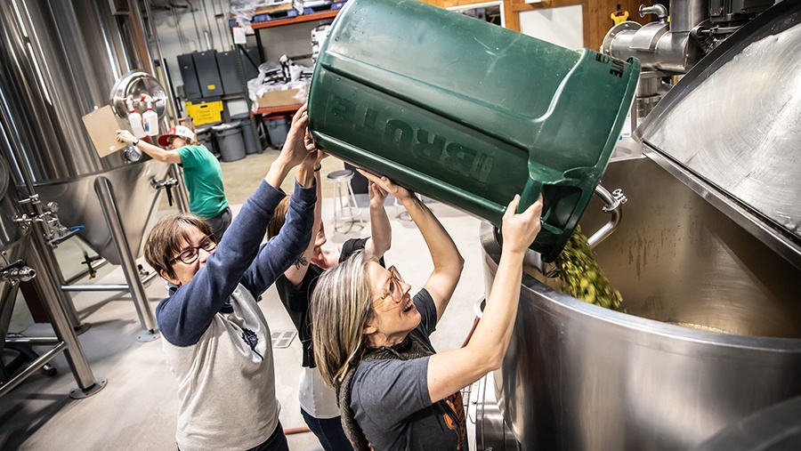 Co-founder Wynne Odell and Merchandise Manager Lynsey Bates pour hops from a green yard bin into a steel fermentation tank used to brew beer