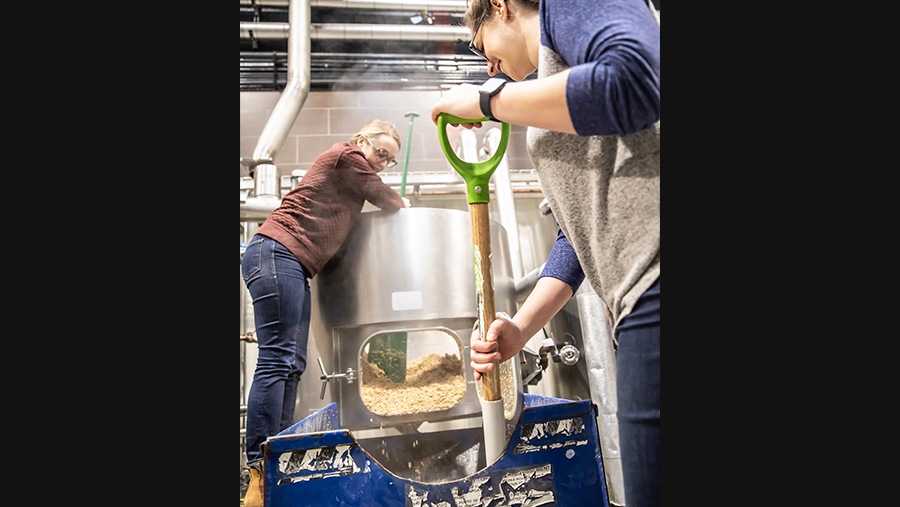 Odell employees Bri Brown and Kristen Wood shovel spent grain out of a steel tank created during the beer making process