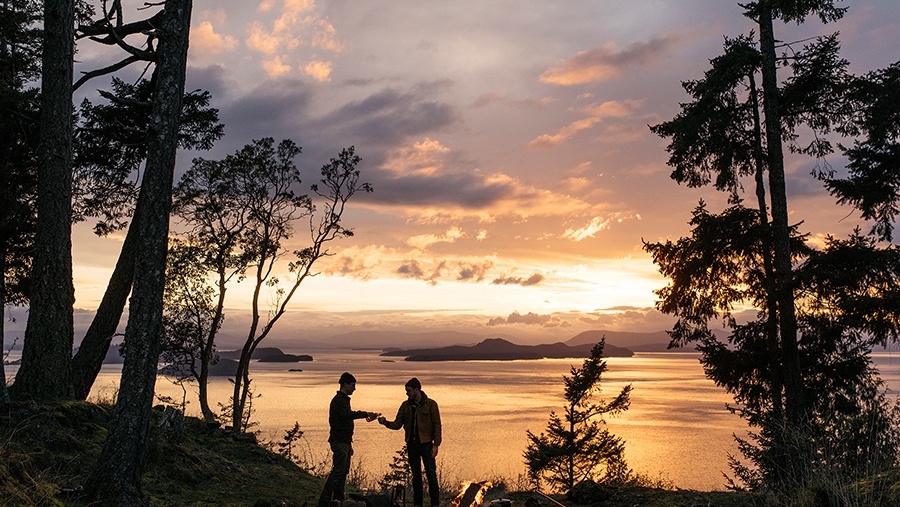 Two friends sharing whiskey near a campfire in a forest on the ocean coast
