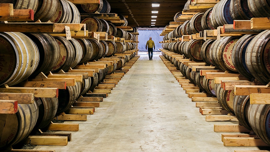 A man in the distance walks between two rows of stacked whiskey barrels in a rackhouse